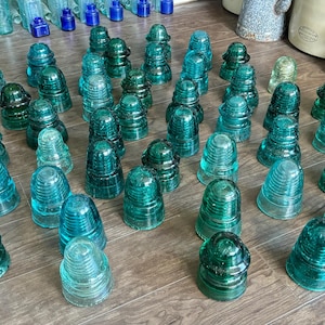 May include: A collection of vintage glass insulators in various shades of green and blue. The insulators are arranged on a wooden surface.
