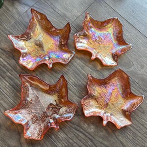 Peut inclure: Un ensemble de quatre coupelles en verre ambré irisé en forme de feuilles d'érable. Chaque coupelle présente une surface texturée avec un motif de nervures prononcé et une finition brillante et réfléchissante aux reflets orange, dorés et violets.