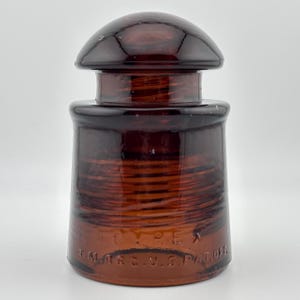 May include: A dark amber glass electrical insulator with a rounded top and a cylindrical body. The glass features embossed text near the base that reads PYREX and T.M. REG. U.S. PAT. OFF. The surface is smooth and translucent with a glossy finish.