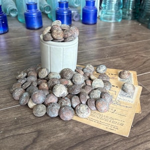 May include: A collection of antique buttons, many with a circular design, spilling from a small, light gray ceramic container. Some buttons are scattered on a vintage paper document. Background includes blue glass bottles.