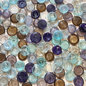 Op de afbeelding: Een verzameling antieke glazen potdeksels in verschillende kleuren, waaronder helder, bruin, blauwgroen en paars. De deksels zijn rond met reliëfontwerpen en texturen, wat een vintage uitstraling creëert. Sommige deksels hebben een verhoogd midden.