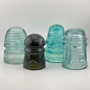May include: Four vintage glass electrical insulators in varying shades of blue, green, and dark olive. Each piece features a rounded top, ribbed sides, and embossed markings including the text CANADIAN and a diamond shape on the glass surface.