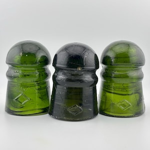 May include: Three antique glass insulators in shades of green and black. The insulators have a rounded top and a diamond-shaped design. The glass has a textured appearance, with visible cracks and imperfections.