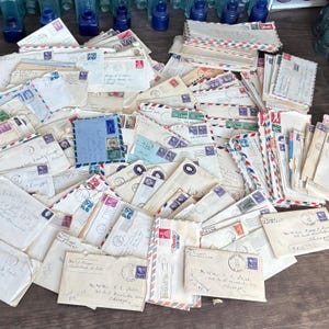 Könnte beinhalten: Eine Sammlung von Vintage-Umschlägen, viele mit Briefmarken und Poststempeln. Die Umschläge sind in verschiedenen Größen und Farben gehalten, einige mit rot-weiß-blauen Streifen. Im Hintergrund sind ein paar blaue Glasflaschen zu sehen.