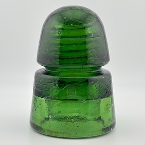 Puede incluir: Un aislante eléctrico de vidrio verde esmeralda vintage con una cúpula superior redondeada y una base cilíndrica más ancha. El material de vidrio translúcido contiene burbujas de aire visibles e imperfecciones internas, con una sección roscada.