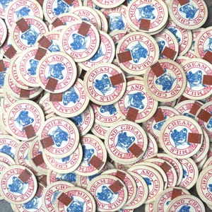 Può includere: Una collezione di tappi di bottiglia di latte e panna vintage. Ogni tappo è rotondo con uno sfondo color crema, testo rosso e blu e una grafica di mucca. Una piccola linguetta marrone rettangolare è attaccata a ogni tappo.