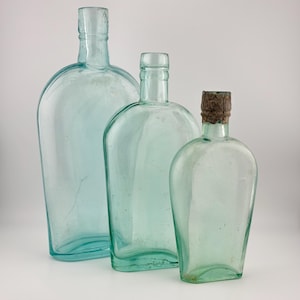 May include: Three clear, light blue-tinted glass bottles of varying heights arranged in a row. Each bottle features a flattened, oval-shaped body with a narrow neck and a rounded rim. The smallest bottle on the right includes a textured cork stopper.