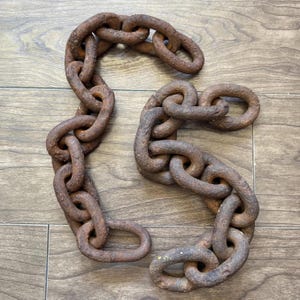 May include: Two sections of a heavy, rusty metal chain are arranged on a wooden surface. The links are large and oval-shaped, showing signs of age and wear. The chains have a dark brown color.