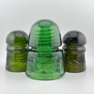 Puede incluir: Tres aisladores eléctricos de vidrio vintage en varios tonos de verde. La pieza central es de un verde esmeralda vibrante, mientras que los dos aisladores laterales son de un verde oliva más oscuro. Cada uno presenta una parte superior redondeada.