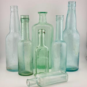 May include: A collection of six vintage glass bottles in varying shades of light green and clear glass. Five bottles stand upright in different heights and shapes, while one rectangular bottle lies horizontally in the foreground.