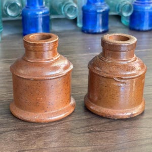 May include: Two brown stoneware inkwells with a cylindrical body, a wide base, and a narrow neck. The inkwells feature a smooth, glazed finish with subtle speckling throughout the clay surface. One inkwell displays a small, embossed letter S on the side.