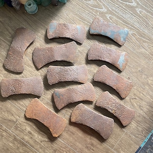 May include: A collection of vintage axe heads with a weathered, rusty appearance. The axe heads are various sizes and shapes, arranged on a wooden surface. The metal has a brown and blue-grey patina, indicating age and use.