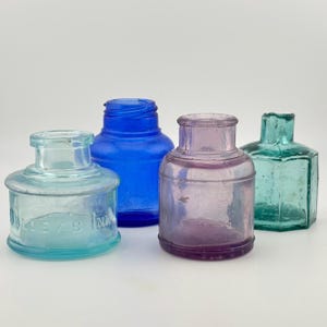 May include: A collection of four glass ink bottles in various colours and shapes. One is light blue with the embossed text "DALLEY'S INK", one is deep cobalt blue, one is a translucent purple, and one is a hexagonal teal bottle with a wide neck.