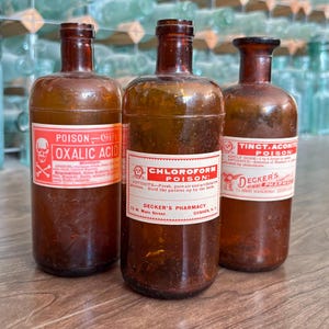 May include: Three vintage amber glass apothecary bottles with narrow necks. Each bottle features a white and red label with black text. The labels read Oxalic Acid, Chloroform Poison, and Tinct. Aconite Poison, all from Decker's Pharmacy in Goshen, NY.
