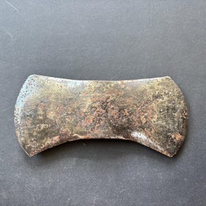 Puede incluir: Una cabeza de hacha de doble filo hecha de metal gris oscuro desgastado con signos visibles de oxidación, óxido y picaduras en la superficie. La herramienta presenta dos filos curvos simétricos a ambos lados de un ojo central.