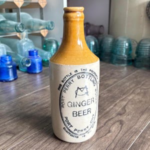 May include: A vintage ceramic ginger beer bottle with a tan base and a brown glazed neck. The front features black printed text reading THIS BOTTLE IS THE PROPERTY OF PORT PERRY BOTTLING WORKS, GINGER BEER, PORT PERRY, ONT, CONTENTS 10 FL OZ.