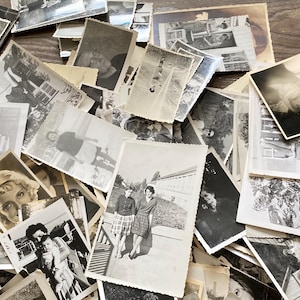 May include: A large pile of black and white photographs, some with faded edges, are stacked on a wooden surface. The photos are of various sizes and shapes, and some have handwritten notes on the back.