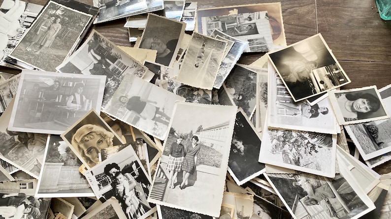 May include: A large pile of black and white photographs, some with faded edges, are stacked on a wooden surface. The photos are of various sizes and shapes, and some have handwritten notes on the back.