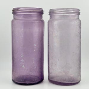 Peut inclure: Deux bocaux cylindriques en verre avec une teinte violette claire. Chaque bocal présente un goulot fileté pour un couvercle à vis, un corps lisse et une base plate. Le verre a un aspect légèrement patiné avec des textures de surface subtiles.