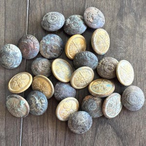 May include: A collection of antique buttons in various shapes and sizes. Some buttons are round with embossed designs, while others are oval with a starburst pattern and the word "CANADA". The buttons have a weathered, aged appearance with shades of bronze, gold, and grey.