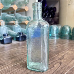 May include: A clear, light green glass bottle with a rectangular body and vertical ribbed sides. The front of the bottle features embossed text that reads POISON and NOT TO BE TAKEN. The bottle has a narrow neck and a rounded, thick glass rim.