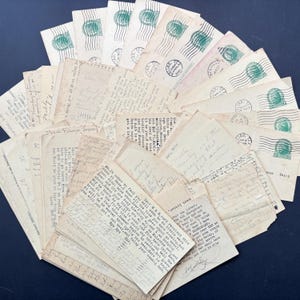 May include: A collection of vintage letters and envelopes, arranged in a fan shape. The envelopes have green postage stamps and postmarks. The handwritten letters are on aged, cream-coloured paper, with visible ink and cursive handwriting. The letters appear to be from the mid-20th century.