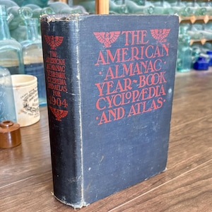 May include: A vintage book titled "The American Almanac Year Book Cyclopedia and Atlas for 1904." The book has a dark blue cover with red lettering and decorative elements. The book is standing upright.