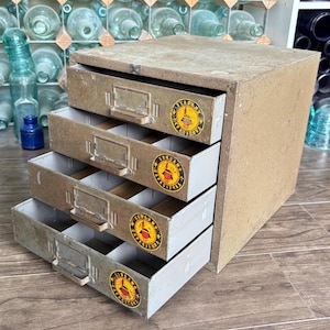 May include: A vintage metal cabinet with four drawers, each labeled with a circular sticker. The cabinet has a weathered, aged appearance, with visible wear and tear. The drawers are open, revealing empty compartments.