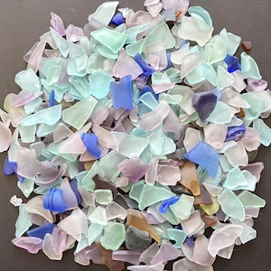 May include: A collection of small, smooth, frosted sea glass pieces in various shapes and sizes. The assortment features a palette of light blue, aqua, pale pink, lavender, white, and deep cobalt blue, with occasional brown and grey fragments.