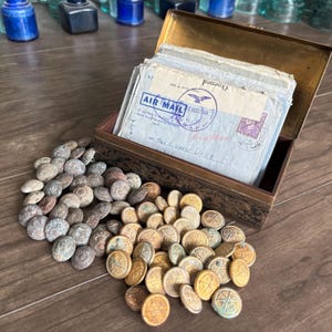 Può includere: Un'antica scatola di ottone aperta piena di lettere aeree vintage, con un mucchio di bottoni antichi davanti. I bottoni sono in due pile, una di bottoni grigi e marroni e l'altra di bottoni color oro.