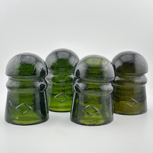 Puede incluir: Cuatro aisladores eléctricos de vidrio verde oscuro con una parte superior redondeada y una base acampanada. Cada pieza presenta una forma de diamante en relieve en la sección inferior, con un acabado liso y translúcido y burbujas de aire visibles.