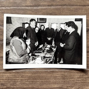 May include: Black and white photograph of a group of people gathered around a table. The table is covered with a patterned cloth. Several people are looking at the table. The photograph is vintage and appears to be from the mid-20th century.