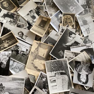 May include: A large pile of black and white photographs, some with faded edges, are stacked on top of each other. The photos are of various sizes and shapes, and some have handwritten notes on the back.