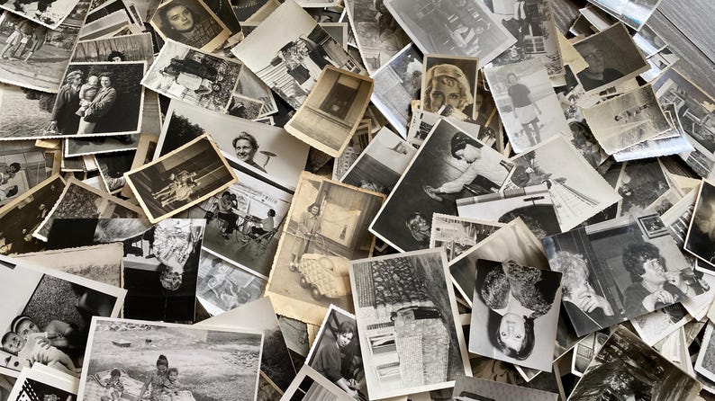 May include: A large pile of black and white photographs, some with faded edges, are stacked on top of each other. The photos are of various sizes and shapes, and some have handwritten notes on the back.