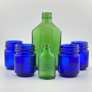 Könnte beinhalten: Eine Sammlung von fünf Glasflaschen, bestehend aus einer großen grünen Flasche, einer kleinen grünen Flasche und drei kobaltblauen Gläsern. Die große grüne Flasche hat einen Gewindehals und geprägten Text auf der Schulter. Die blauen Gläser haben Gewinderänder.