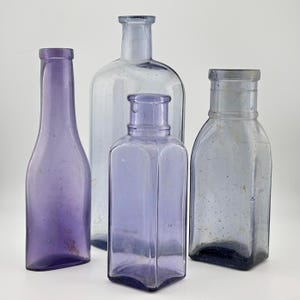 Peut inclure: Quatre bouteilles en verre antiques de couleurs violettes et transparentes. Les bouteilles varient en forme et en taille, avec une bouteille haute et élancée et une bouteille carrée à base hexagonale. Les bouteilles sont présentées sur un fond blanc uni.