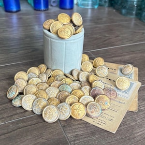 May include: A collection of vintage gold-toned buttons, many with a Canada emblem, spilling from a small, light-colored ceramic container. Some buttons are scattered on a wooden surface, alongside aged paper documents.