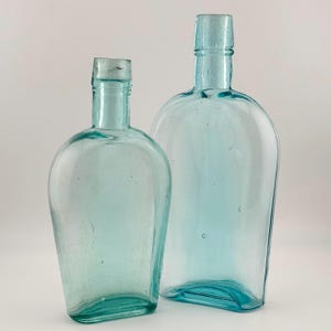 May include: Two vintage-style glass bottles in a light aqua blue hue. The bottles feature a rounded, flattened flask shape with narrow necks and clear glass construction, displaying subtle air bubbles and variations within the glass material.