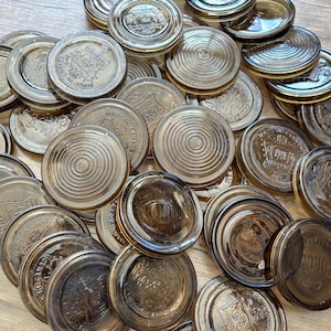 Puede incluir: Una colección de tapas de tarros de vidrio ámbar vintage. Las tapas presentan diseños en relieve, incluyendo círculos concéntricos y texto como "Made in Canada". Las tapas están dispuestas sobre una superficie de madera.