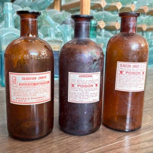 May include: Three vintage amber glass apothecary bottles with narrow necks and wide bases. Each bottle features a white paper label with red text and a skull and crossbones symbol. The labels are marked Chloroform Liniment and Ammonia.