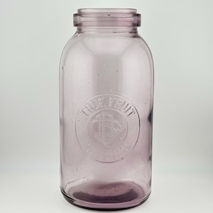 Peut inclure: Un bocal en verre violet clair avec une large ouverture et un bord épais. L'avant du bocal présente un logo circulaire en relief avec le texte TRUE FRUIT et un motif graphique central. Le verre a une finition lisse et translucide.
