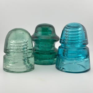 Puede incluir: Tres aisladores eléctricos de vidrio vintage en varios tonos de verde y azul. El aislador izquierdo es verde claro, el central es verde oscuro con el texto BROOKFIELD y el derecho es azul brillante con una estrella en relieve en la base.
