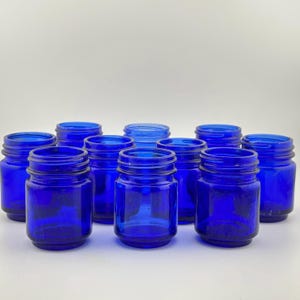 Könnte beinhalten: Eine Ansammlung von zehn kleinen, leeren Gläsern aus kobaltblauem Glas, die in einer Gruppe angeordnet sind. Jedes Glas hat einen zylindrischen Körper mit einem Gewinderand an der Oberseite, der für einen Schraubdeckel vorgesehen ist.