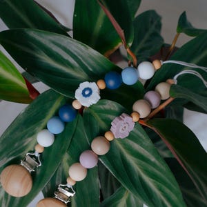 May include: Two wooden baby pacifier clips with colorful beads and flower accents. One clip features blue, white, and wooden beads, with a white flower. The other clip has pink, beige, and wooden beads, with a lavender flower. The clips have wooden bases and metal clasps.