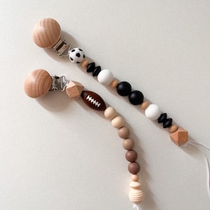 Op de afbeelding: Twee houten baby-bijtjes met clips. Eén bijtje heeft een voetbalbedel en bruine, beige en crème kralen. De andere bijtje heeft een voetbalbedel en zwarte, witte en bruine kralen.