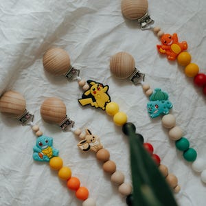 Op de afbeelding: Een verzameling fopspenen met houten kralen en kleurrijke siliconen bedels. De bedels tonen Pikachu, Charmander, Squirtle, Bulbasaur en Eevee. De clips hebben een houten clip en liggen op een witte ondergrond.