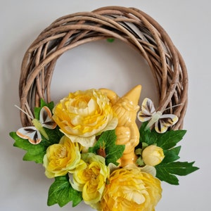Pode incluir: Uma grinalda decorativa com uma base de videira tecida. É adornada com flores amarelas vibrantes, folhas verdes, uma figura de gnomo amarelo e dois detalhes de borboleta. Um charmoso item de decoração para casa na primavera ou verão.