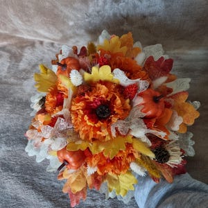 Peut inclure: Un bouquet de fleurs d'automne avec des fleurs orange, jaunes et rouges, des feuilles et de petites citrouilles. Le bouquet est lié avec un ruban blanc et a une ambiance rustique et automnale.