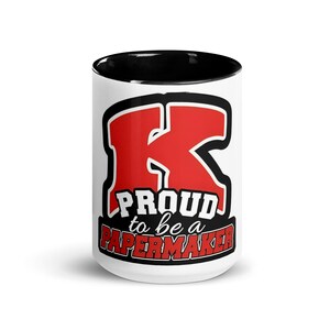 May include: White ceramic mug with a black rim. The mug features a red and black graphic of a large letter "K" with the text "Proud to be a Papermaker".