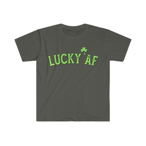 Könnte beinhalten: Ein dunkelgraues T-Shirt mit einem grünen Kleeblatt und dem Text "LUCKY AF" in grün.